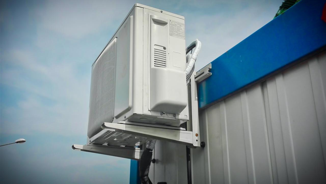 Zewnętrzna jednostka klimatyzatora zamontowana na metalowej konstrukcji przy budynku z blachy trapezowej, widok z dołu na tle błękitnego nieba.