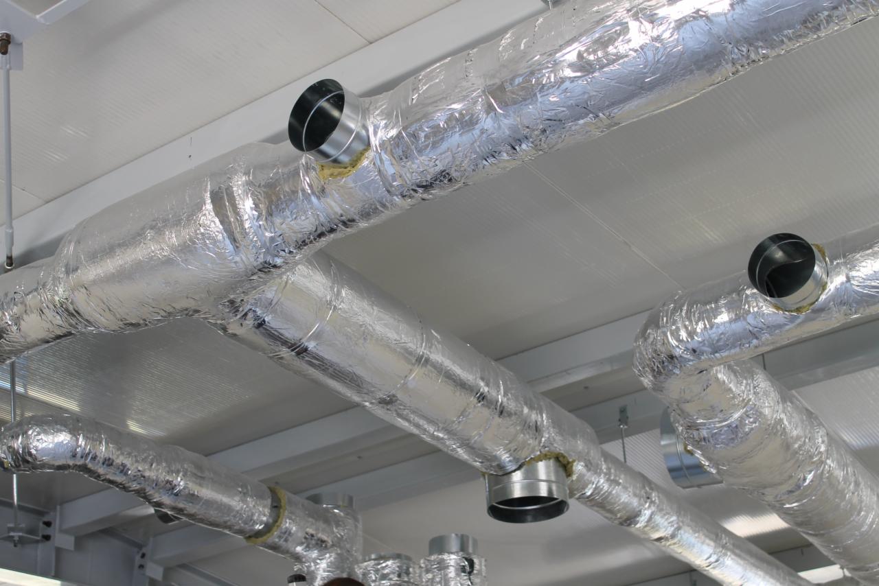 Srebrne, izolowane przewody wentylacyjne zamontowane pod sufitem; widoczne metalowe końcówki i łączenia. Instalacja w jasnym pomieszczeniu.