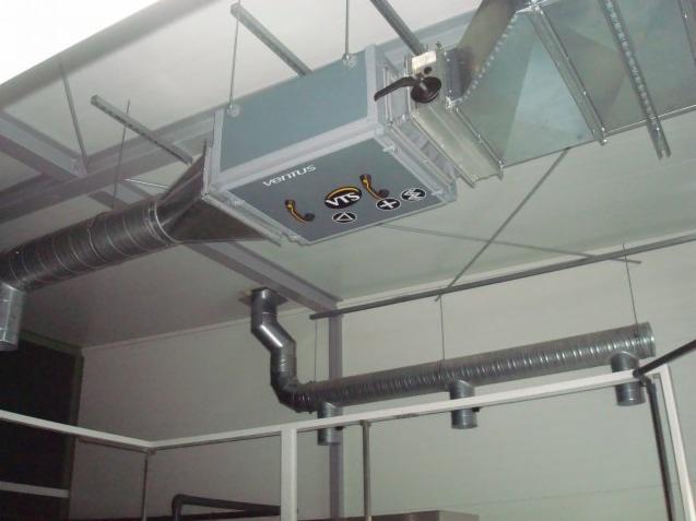 Szary centralny element wentylacyjny marki VTS, zamontowany pod sufitem, z podłączonymi metalowymi rurami wentylacyjnymi o przekroju okrągłym, widoczne elementy konstrukcji wsporczej