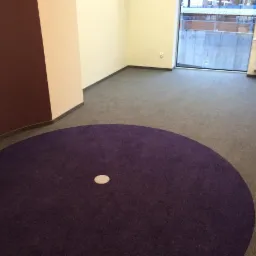 Szara wykładzina w biurze z okrągłym, fioletowym dywanem i widocznym włazem rewizyjnym. Ściany w odcieniach beżu i bordo. W tle okno i grzejnik.