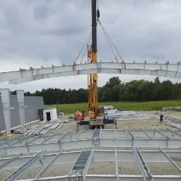 Montaż stalowej konstrukcji hali przemysłowej za pomocą dźwigu, widoczne elementy szkieletu i przygotowane profile, w tle zieleń i pochmurne niebo.