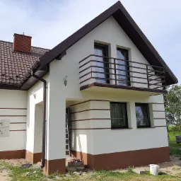 Dwukondygnacyjny dom z balkonem, świeżo pomalowaną elewacją w kolorze białym z brązowymi akcentami, brązowy dach, widoczna drabina i narzędzia sugerujące prace wykończeniowe.