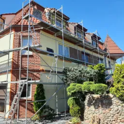 Dwukondygnacyjny dom z ceglaną elewacją i fragmentami ocieplenia, otoczony rusztowaniem, z widocznym pracownikiem na dachu oraz ozdobnym ogrodem skalnym