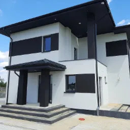 Dwukondygnacyjny dom z białą elewacją i czarnymi akcentami, widok z zewnątrz na wejście z schodami, teren w trakcie prac wykończeniowych.