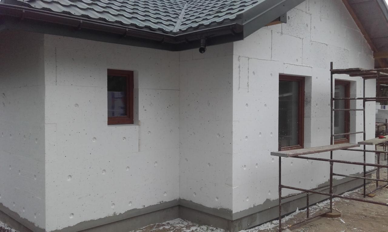 Dom w trakcie ocieplania: białe płyty styropianowe z widocznymi otworami montażowymi, brązowe ramy okienne, szare elementy dachu i rusztowanie przy ścianie.