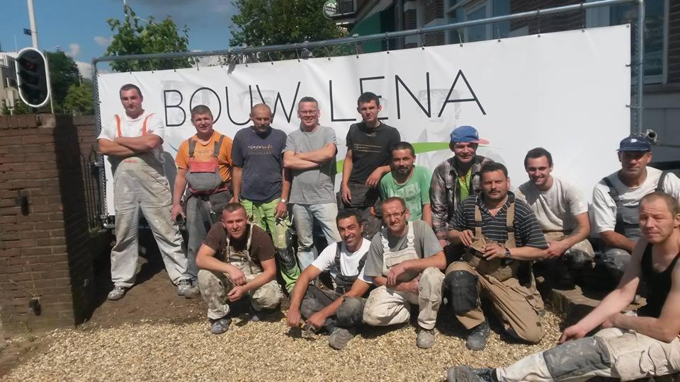 Grupa trzynastu pracowników budowlanych w ubraniach roboczych pozuje na tle banneru z nazwą firmy 'BOUWLENA', stojącego na żwirowym podłożu, widoczne budynki w tle.