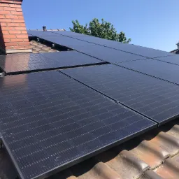 Panele słoneczne zamontowane na dachu z ceglaną dachówką, widoczny komin w tle, błękitne niebo.