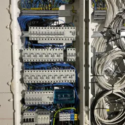 Rozdzielnica elektryczna z zabezpieczeniami, bezpiecznikami i przewodami w kolorach niebieskim, czarnym i białym, zamontowana w surowej ścianie z widoczną pianką montażową, widok z bliska.