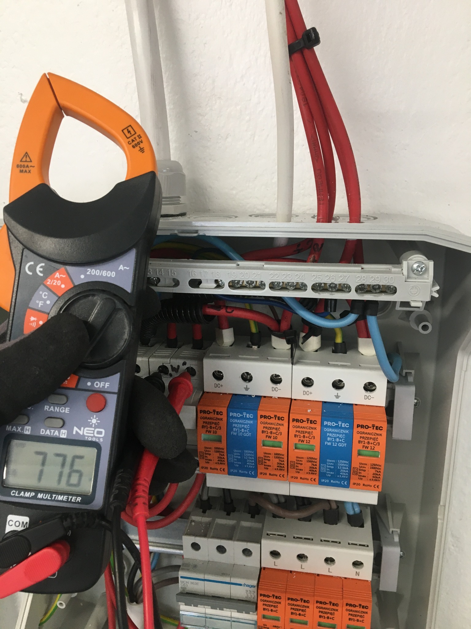 Elektryk w czarnych rękawicach ochronnych mierzy napięcie w rozdzielnicy z zabezpieczeniami przepięciowymi Pro-Tec, wskazanie multimetru Neo Tools to 776.