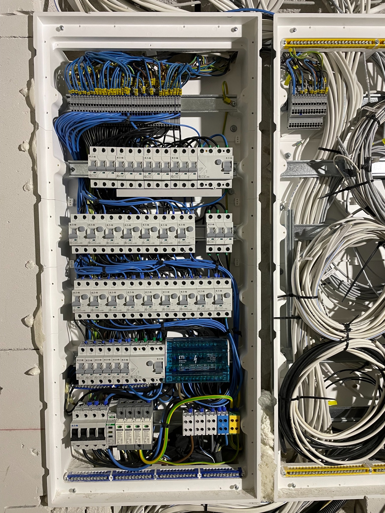 Rozdzielnica elektryczna z zabezpieczeniami, bezpiecznikami i przewodami w kolorach niebieskim, czarnym i białym, zamontowana w surowej ścianie z widoczną pianką montażową, widok z bliska.