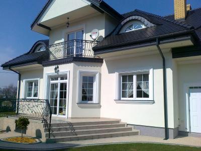 Elewacja domu z balkonem i schodami wejściowymi, biała fasada, czarny dach, eleganckie detale architektoniczne, zadbany trawnik, słoneczny dzień.