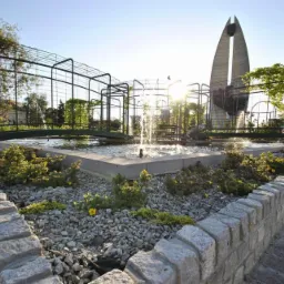 Oczko wodne z fontanną i metalową konstrukcją porośniętą roślinnością, otoczone kamiennym murkiem i brukowaną ścieżką, w tle nowoczesny pomnik i budynki.