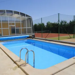 Prostokątny basen z niebieską wodą, metalowymi schodkami i częściowo zwiniętą, niebieską pokrywą, osłonięty przeszkloną konstrukcją, z kortem tenisowym i boiskiem do koszykówki w tle.
