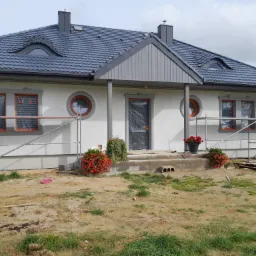 Dom jednorodzinny w trakcie prac elewacyjnych; widoczne rusztowania, okna zabezpieczone folią, świeżo położona jasna elewacja, dekoracyjne okrągłe okna, ciemna dachówka.