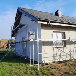 Dom jednorodzinny w trakcie tynkowania elewacji, z widocznym rusztowaniem, zabezpieczonymi oknami folią i fragmentem placu zabaw w tle.
