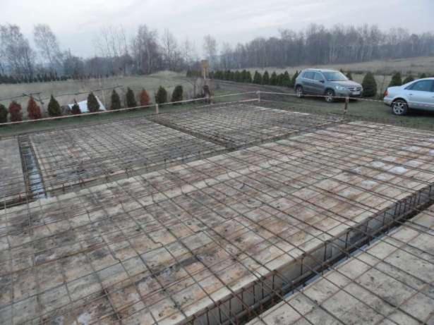Zbrojenie fundamentów domu jednorodzinnego, widok z góry. Metalowa siatka na betonowych podkładach, w tle zieleń i dwa samochody. Pochmurny dzień.