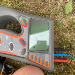 Miernik Sonel MRU-20 na trawie, z podłączonymi przewodami pomiarowymi (czerwony, niebieski, czarny), wskazujący wynik pomiaru rezystancji uziemienia 137 omów, widoczny fragment nogi operatora...
