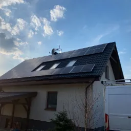 Dom jednorodzinny z zamontowanymi panelami słonecznymi na dachu, widoczny biały samochód dostawczy z bagażnikiem dachowym i anteną satelitarną na dachu, błękitne niebo z chmurami w tle, ujęcie z dołu.