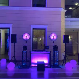 Scena z konsoletą DJ-ską, nagłośnieniem i oświetleniem w fioletowej tonacji, ustawiona w eleganckim wnętrzu z kolumnami i szklanymi drzwiami.