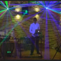 DJ obsługuje konsoletę z laptopem, w tle ceglana ściana oświetlona kolorowymi laserami i reflektorami.