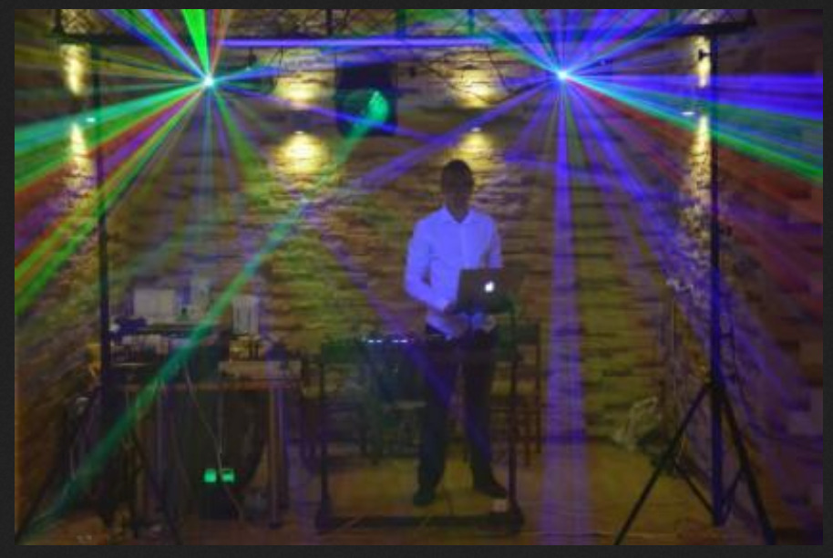 DJ obsługuje konsoletę z laptopem, w tle ceglana ściana oświetlona kolorowymi laserami i reflektorami.