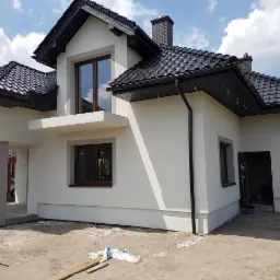 Biały dom jednorodzinny z ciemną dachówką i stolarką okienną. Elewacja świeżo po tynkowaniu, widoczne rynny i elementy wykończeniowe. Dom w stylu nowoczesnym, z wyraźnym zarysem bryły.