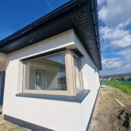 Fragment nowo otynkowanego domu jednorodzinnego z oknem o drewnianej ramie i czarnym wykończeniem dachu, widok z zewnątrz na tle błękitnego nieba z chmurami.