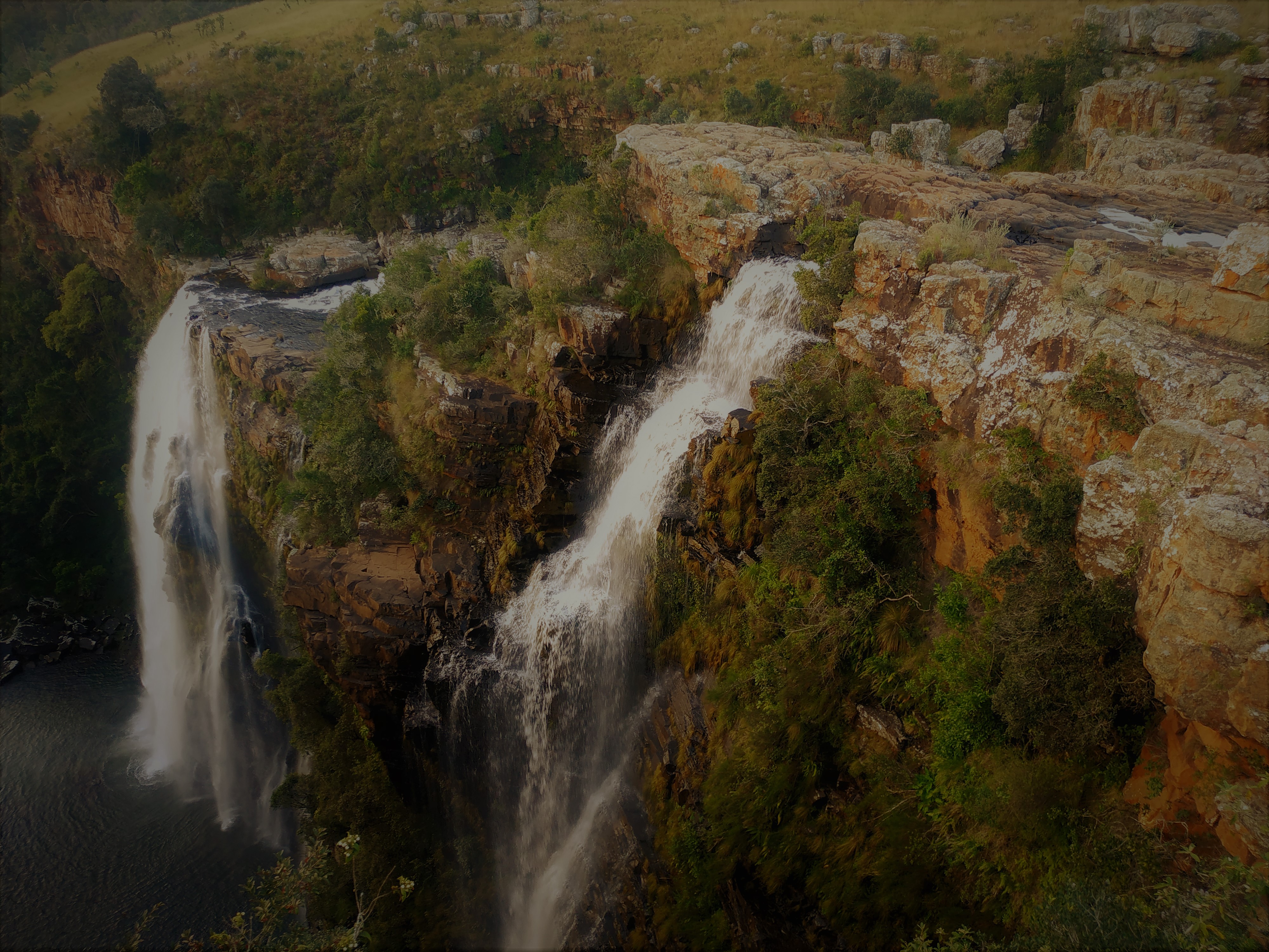 Panorama wodospadu z kaskadami spływającymi po skalistym klifie porośniętym zielenią, w tle wzgórza i jasne niebo.