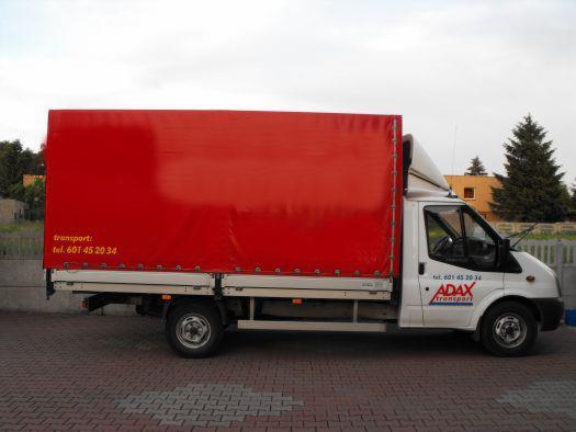 Biała furgonetka z czerwoną plandeką i logo Adax Transport parkuje na brukowanej ulicy. Widok z boku, drzewa w tle.