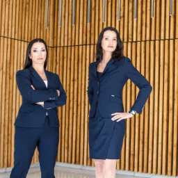 Dwie pewne siebie prawniczki w granatowych garniturach pozują na tle drewnianej ściany w nowoczesnym biurze, jedna z założonymi rękami, druga z ręką na biodrze.