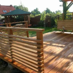 Drewniany taras z balustradą z poziomych desek, w tle altana ogrodowa i dom z czerwonym dachem. Jasne drewno, słoneczny dzień.