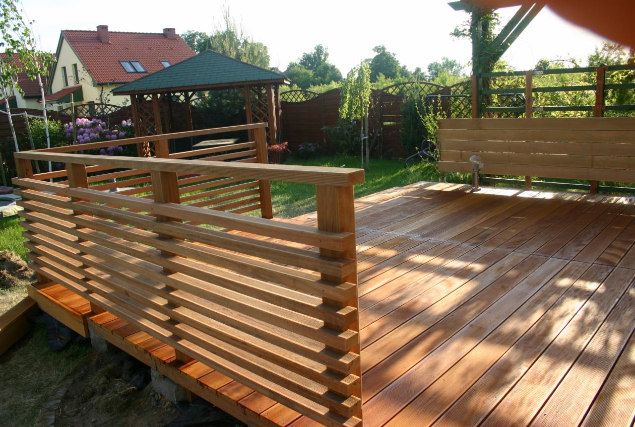 Drewniany taras z balustradą z poziomych desek, w tle altana ogrodowa i dom z czerwonym dachem. Jasne drewno, słoneczny dzień.