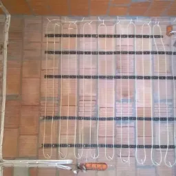 Montaż ogrzewania podłogowego na ceglanej ścianie. Rury grzewcze przymocowane do czarnych listew. Widoczne przewody elektryczne i narzędzia.