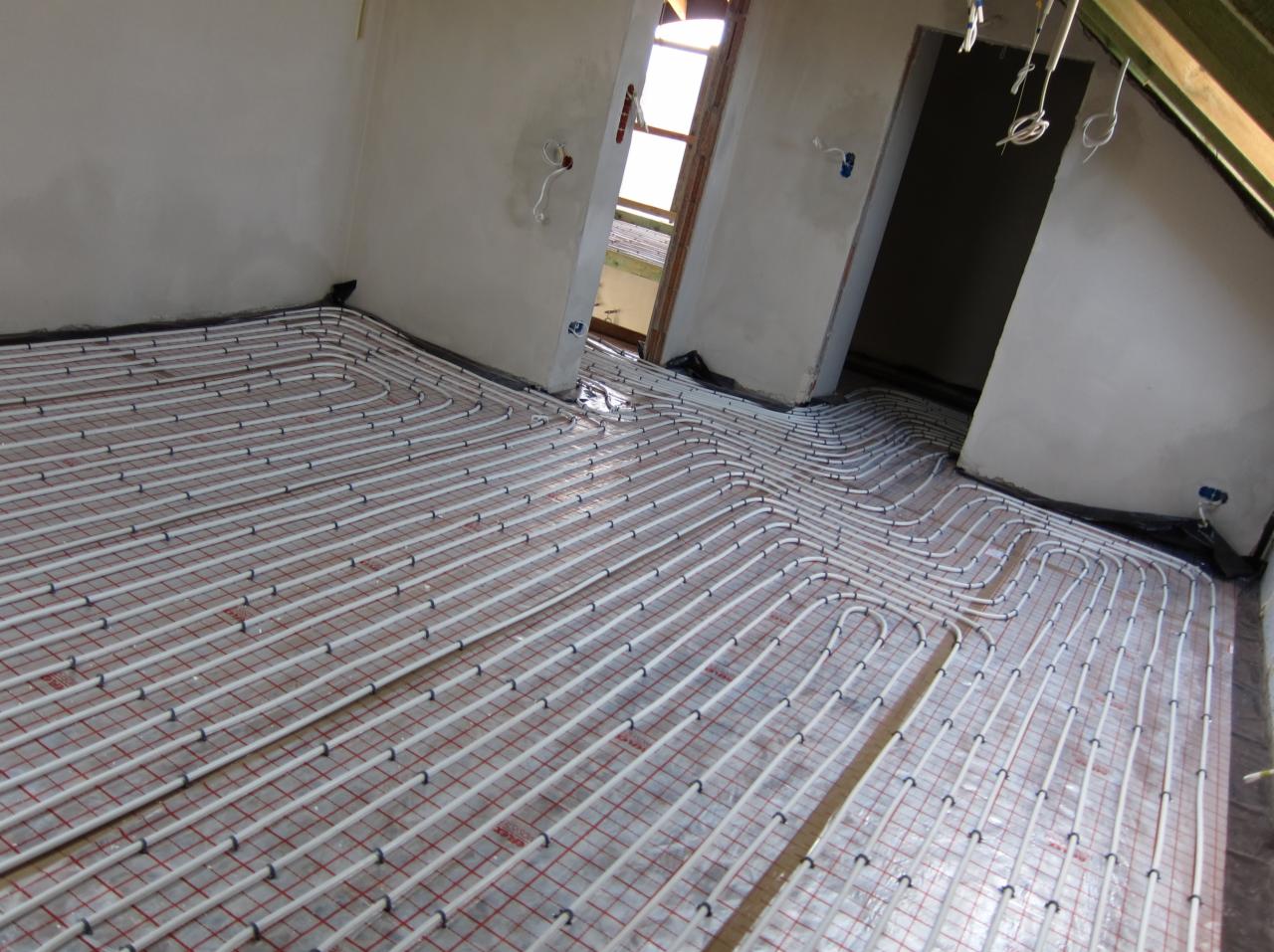Białe rury ogrzewania podłogowego ułożone na czerwonej siatce w surowym wnętrzu poddasza, widoczne fragmenty ścian i okien. Etap instalacji przed wylewką.