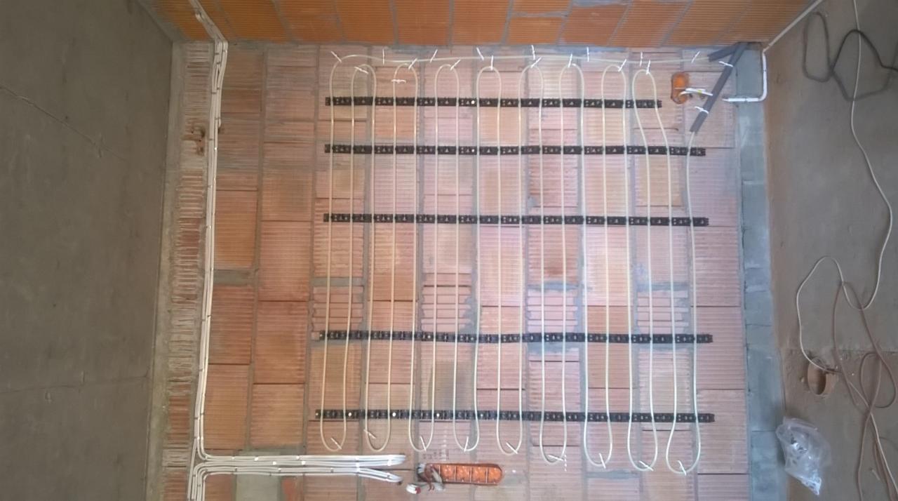 Montaż ogrzewania podłogowego na ceglanej ścianie. Rury grzewcze przymocowane do czarnych listew. Widoczne przewody elektryczne i narzędzia.