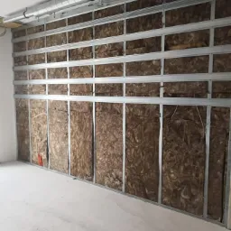 Ściana z metalowym stelażem wypełnionym brązową wełną mineralną, widoczna instalacja wentylacyjna pod sufitem, surowy stan wykończenia wnętrza.