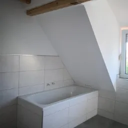 Wykończona łazienka na poddaszu z białą wanną obudowaną płytkami, szarą podłogą i drewnianymi belkami stropowymi.