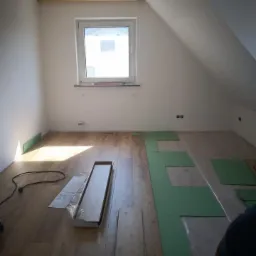 Montaż paneli podłogowych w pokoju na poddaszu ze skosem, widoczna rozłożona zielona pianka podkładowa i paczka paneli w opakowaniu.