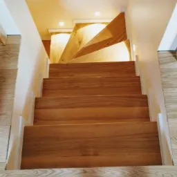 Jasne, drewniane schody z równymi stopniami, widziane z góry, z delikatnym oświetleniem punktowym. Widoczne elementy wykończenia w odcieniach bieli i beżu.