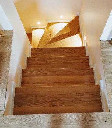 Jasne, drewniane schody z równymi stopniami, widziane z góry, z delikatnym oświetleniem punktowym. Widoczne elementy wykończenia w odcieniach bieli i beżu.