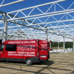 Konstrukcja stalowa hali w trakcie budowy, widoczny czerwony bus firmy Stalex z napisem „Konstrukcje Stalowe” na tle błękitnego nieba. Słupy i kratownica w jasnym kolorze.