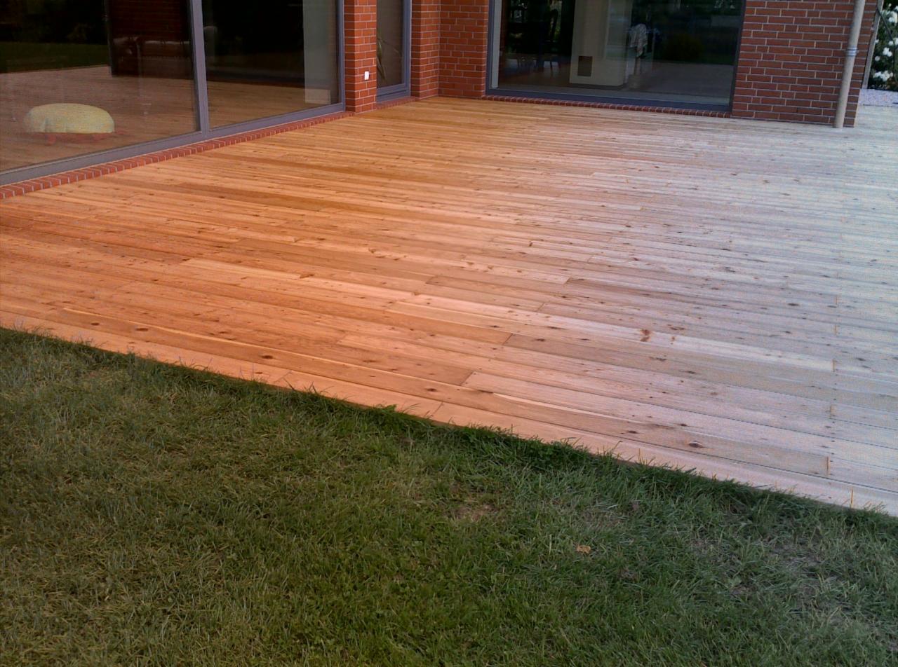 Jasna, drewniana deska tarasowa przylegająca do trawnika, z widoczną ceglaną elewacją budynku w tle i dużymi oknami.