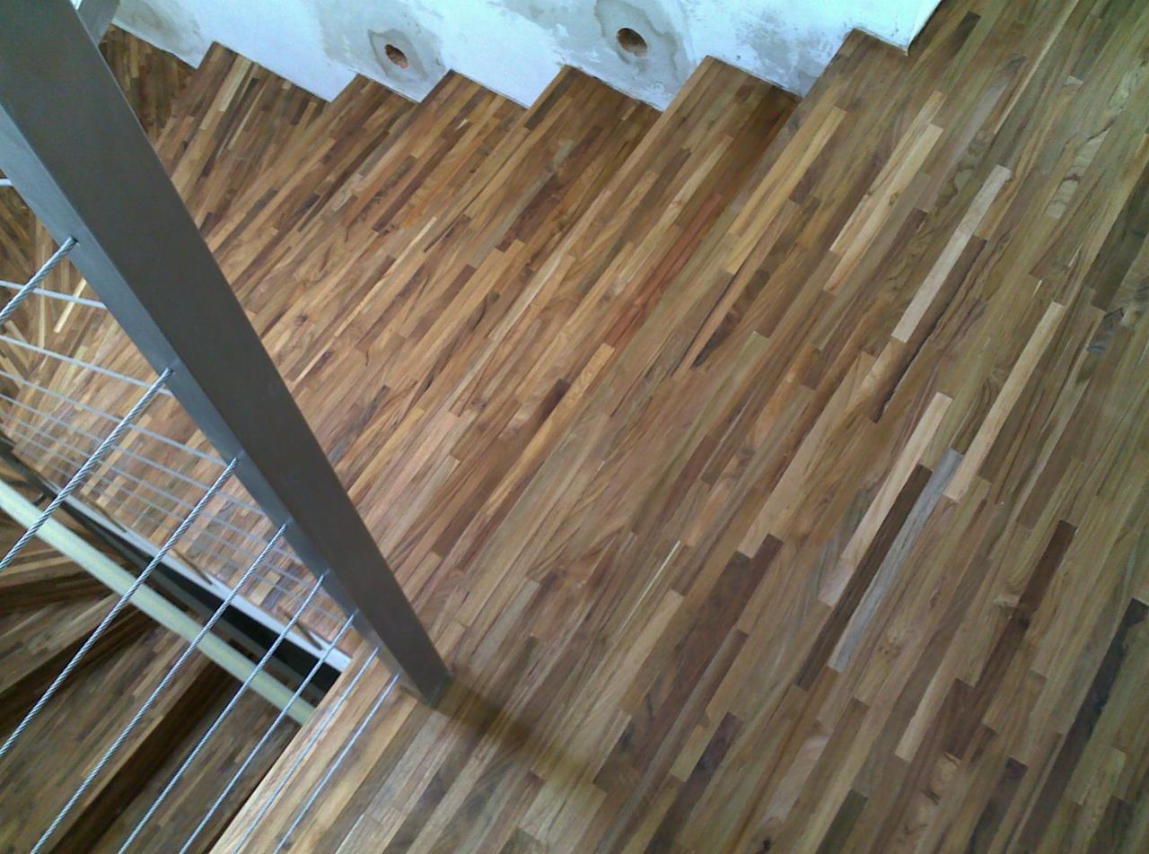 Kątowe ujęcie schodów drewnianych z metalową barierką i linkami. Widoczne wykończenie stopni z różnych odcieni drewna. Ściana w górnej części kadru z widocznymi niedoskonałościami.