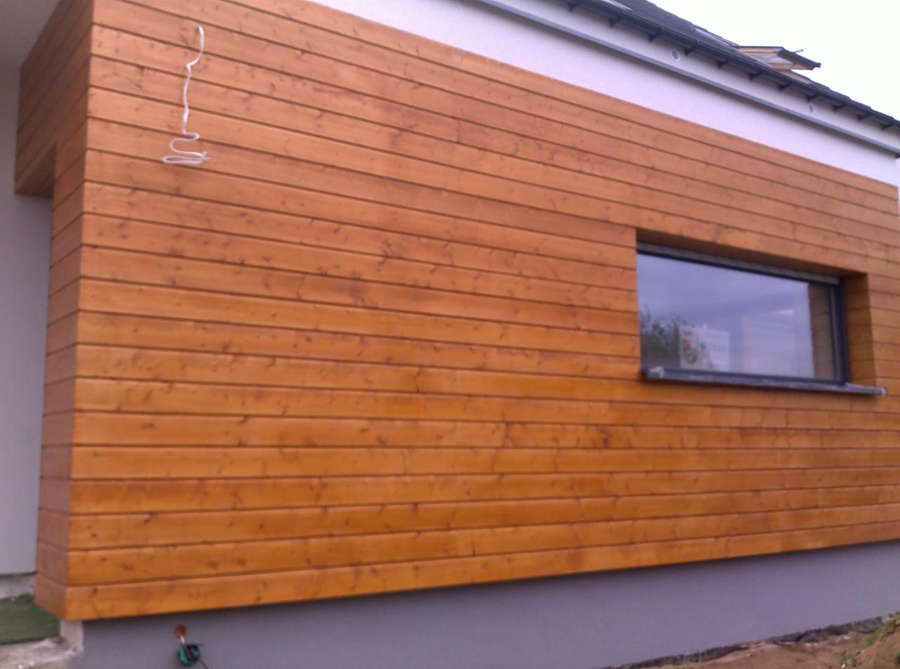 Elewacja domu wykończona poziomymi deskami z drewna w ciepłym, brązowym odcieniu. W ścianie osadzone podłużne okno z czarną ramą. Widoczny fragment dachu i białe wykończenie.