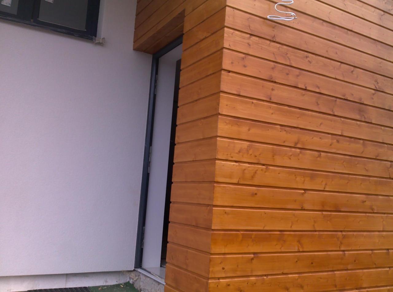 Fragment elewacji budynku z drewnianą okładziną w ciepłym, brązowym odcieniu, widoczna część okna i drzwi. Nowoczesne wykończenie.