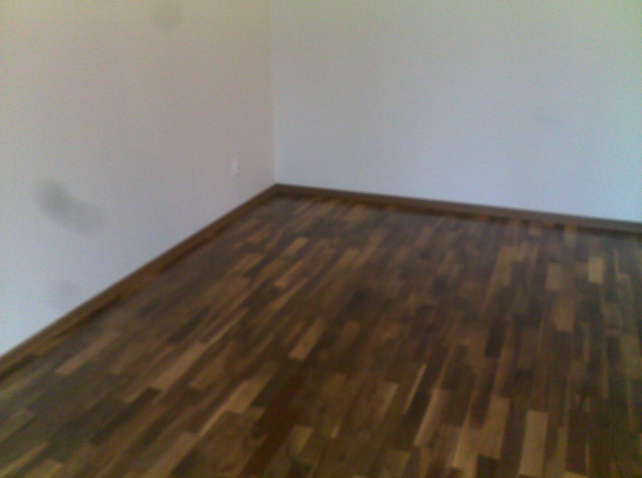 Wykończona podłoga z ciemnego drewna parkietowego w rogu pokoju z białymi ścianami i listwami przypodłogowymi. Widok z góry, naturalne światło.