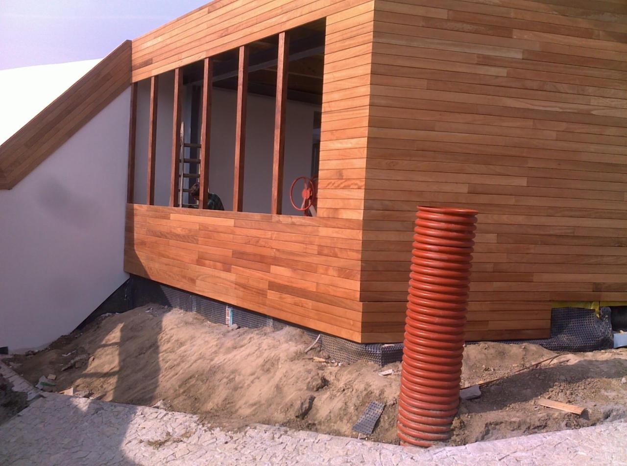 Elewacja budynku wykończona drewnianą boazerią, widoczna pomarańczowa rura kanalizacyjna. Na balkonie drabina i osoba. Częściowo widoczny teren budowy.