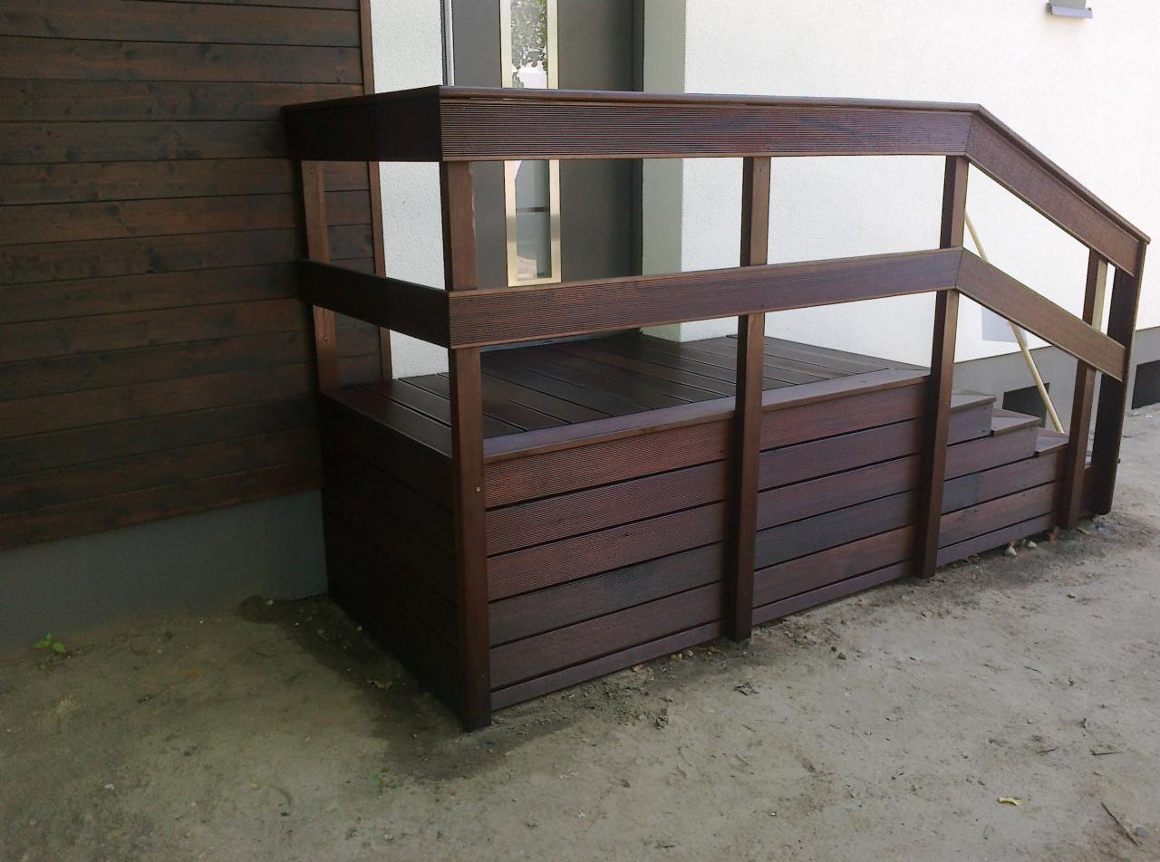 Brunatne schody zewnętrzne z kompozytu, z barierkami, przylegające do budynku z drewnianą elewacją. Widoczny fragment drzwi wejściowych.