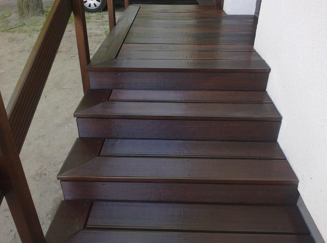 Ciemnobrązowe schody drewniane z ryflowanymi deskami, prowadzące do wejścia. Widoczne elementy balustrady i fragment ściany.