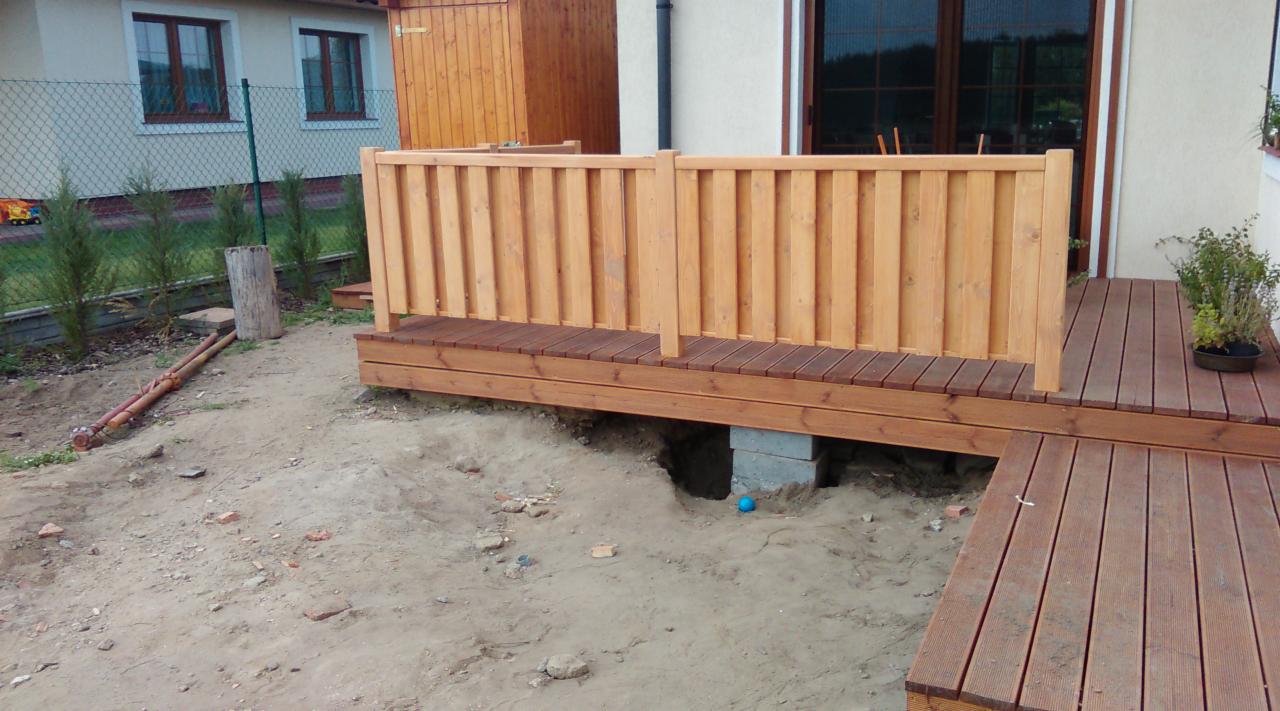 Drewniany taras w trakcie budowy, widoczne deski, barierki z jasnego drewna i betonowe podpory. W tle dom z oknami i zielony ogród.